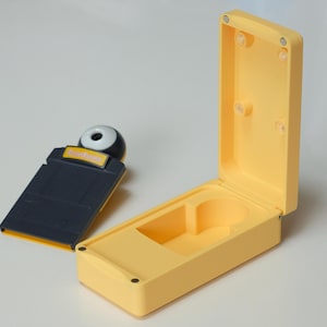 Può includere: Una custodia rettangolare gialla con coperchio aperto e un accessorio nero e giallo separato. L'accessorio ha una parte superiore a forma di sfera bianca e nera e un'etichetta gialla. La custodia è aperta, rivelando un vano interno.