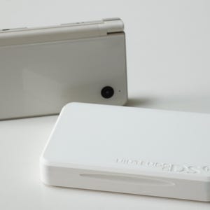 May include: A white Nintendo 3DS handheld game console, closed, with a second white rectangular case in the foreground. The 3DS is in the background, slightly out of focus. The case has the words "Nintendo 3DS" embossed on the top.