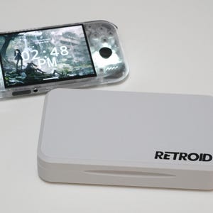 May include: A handheld gaming console with a transparent shell and a screen displaying 02:48 PM, positioned next to a rectangular white storage case featuring the black text RETROID printed on the lower right corner of the lid.