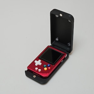 Puede incluir: Una consola de juegos portátil roja con una cubierta abatible negra. El dispositivo cuenta con una pantalla, una cruceta direccional y botones de acción de colores. La cubierta está abierta, revelando los controles.