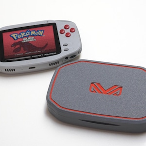 Op de afbeelding: Een grijze draagbare videogameconsole met een scherm dat Pokémon Ruby Version weergeeft, voorzien van een zwarte richtingsknop en rode actieknoppen. Daarnaast ligt een bijpassende grijze rechthoekige opbergdoos met een rode rand.