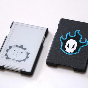 May include: Two black protective cases for an e-reader. One case features a cartoon lion design, and the other has a skull emblem with blue flames. The cases are designed to fit an e-reader device.