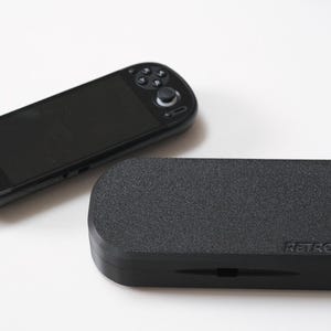 May include: A black handheld games console and a matching black case. The console features a screen, buttons, and control sticks. The case is rectangular with rounded edges and the word "RETROID" printed on it.