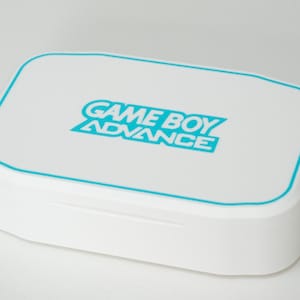 May include: A white Game Boy Advance case with a teal border and teal text. The text reads "GAME BOY ADVANCE" in a stylized font. The case is rectangular with rounded corners.