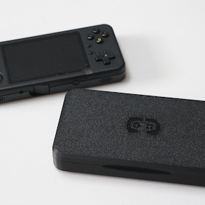 Puede incluir: Una consola de juegos portátil negra con pantalla, cruceta y botones, junto a una funda negra texturizada con un logotipo. La consola y la funda están sobre una superficie blanca. La consola está diseñada para juegos portátiles.