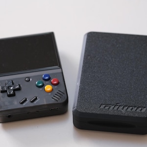 Puede incluir: Una consola de juegos portátil negra con pantalla, cruceta y botones de colores. Al lado, una funda negra con superficie texturizada. La consola tiene un diseño elegante y moderno.