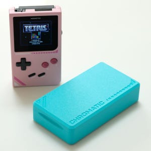 Peut inclure: Une console de jeu portable rose affichant Tetris à l'écran. À côté, une boîte rectangulaire turquoise avec le mot "CHROMATIC" imprimé. La console a une croix directionnelle noire et des boutons roses.