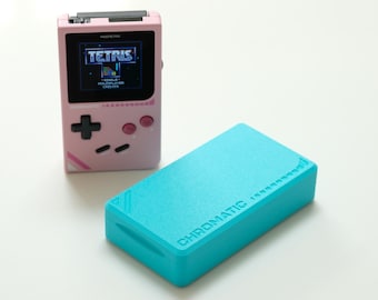 May include: A pink handheld gaming device with the game Tetris displayed on the screen. A turquoise rectangular box with the word "CHROMATIC" is next to the device. The game console has a black directional pad and pink buttons.