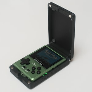 Pode incluir: Um console de jogos portátil verde com uma capa protetora preta. A tela exibe um jogo Pokémon. O dispositivo possui um direcional, botões e o texto "GKD PIXEL" é visível na capa.