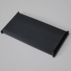 May include: A rectangular, matte black box with slightly raised edges. The box is a solid colour and appears to be made of a textured material. The box is resting on a light grey surface.
