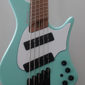 May include: A mint green electric bass guitar with a white pickguard and black pickups. The guitar has five strings and tuning knobs. The brand name "Ibanez" is visible on the headstock.