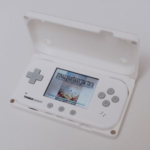 May include: A white handheld gaming console with a screen displaying the game "Final Fantasy Tactics." The console has a gray directional pad, gray buttons, and the text "TRIMUI SMART" is visible.