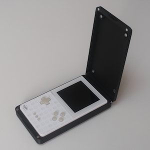 Könnte beinhalten: Ein tragbares Spielgerät mit einem schwarzen Klappgehäuse und einem weißen Innenraum. Das Gerät verfügt über einen Bildschirm, ein Steuerkreuz und mehrere Tasten. Das Gehäuse ist geöffnet und zeigt den Bildschirm und die Bedienelemente. Das Gerät befindet sich auf einer weißen Oberfläche.