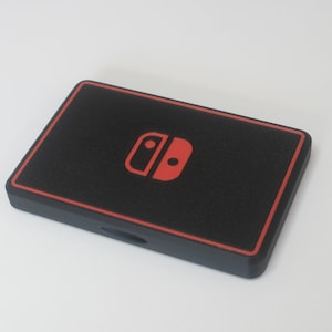 Può includere: Una custodia rettangolare nera con bordo rosso e icona rossa del controller di gioco. La custodia ha una superficie strutturata e un piccolo ritaglio su un lato. Probabilmente è una custodia protettiva per una console di gioco.