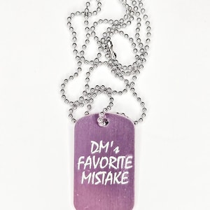 May include: A purple dog tag necklace with the text "DM's FAVORITE MISTAKE" in white. The tag is rectangular with rounded corners and hangs from a silver ball chain necklace. The background is white.