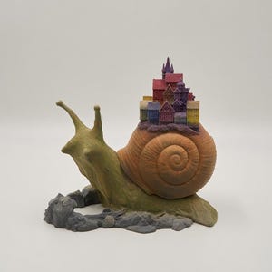 May include: A sculpture of a green snail with a large, textured brown shell. Perched atop the shell is a cluster of miniature, multi-colored buildings in shades of red, purple, blue, and yellow. The snail rests on a base of gray, rocky terrain.