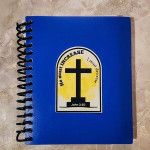 May include: A blue spiral notebook with a sticker featuring a yellow and white watercolor background with a black cross and the text "He must increase, I must decrease" and "John 3:30".