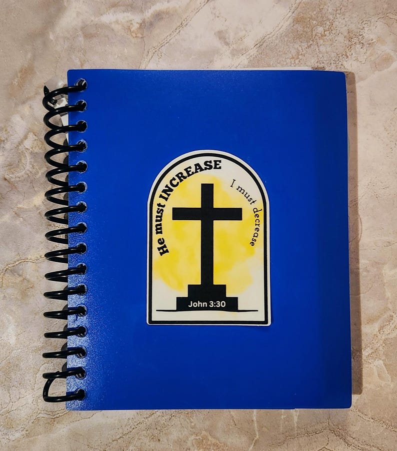 May include: A blue spiral notebook with a sticker featuring a yellow and white watercolor background with a black cross and the text "He must increase, I must decrease" and "John 3:30".