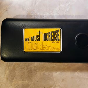 May include: Black power bank with a yellow sticker that reads "He must increase I must decrease John 3:30".