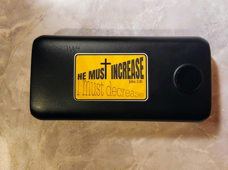 May include: Black power bank with a yellow sticker that reads "He must increase I must decrease John 3:30".