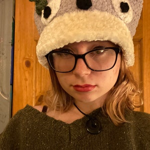 May include: A person wearing a purple and white crocheted hat with black eyes and a green leaf on top. The hat is shaped like a frog. The person is wearing a green sweater and a black necklace with a round pendant.