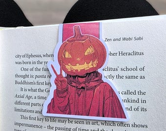 Könnte beinhalten: Lesezeichen mit einer Cartoon-Illustration einer Person mit einem Kürbiskopf und einem roten Kapuzenpullover, die ein Friedenszeichen gibt. Das Lesezeichen befindet sich auf einem Buch mit Text über Zen und Wabi Sabi.