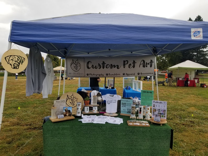 Pode incluir: Uma banca em um mercado ao ar livre vendendo arte personalizada para animais de estima&ccedil;&atilde;o. A banca tem um toldo azul e uma placa que diz "Custom Pet Art" com as palavras "Photography, Tumblers, Apparel, Accessories" listadas abaixo. H&aacute; v&aacute;rios itens &agrave; venda, incluindo canecas, camisetas e retratos emoldurados.