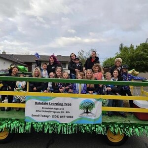 Pode incluir: Um grupo de crian&ccedil;as e adultos est&atilde;o sentados em um carro aleg&oacute;rico verde e amarelo decorado com guirlandas verdes. O carro aleg&oacute;rico tem uma placa que diz "Oakdale Learning Tree" e inclui informa&ccedil;&otilde;es de contato.
