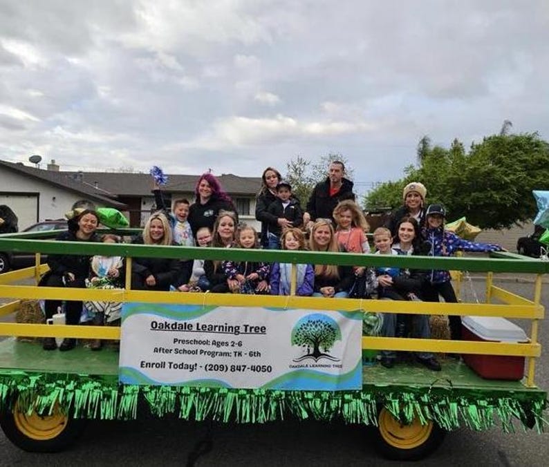 Pode incluir: Um grupo de crian&ccedil;as e adultos est&atilde;o sentados em um carro aleg&oacute;rico verde e amarelo decorado com guirlandas verdes. O carro aleg&oacute;rico tem uma placa que diz "Oakdale Learning Tree" e inclui informa&ccedil;&otilde;es de contato.