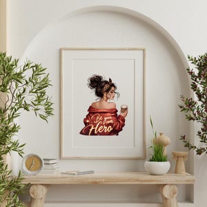 May include: Framed art print featuring a woman with dark hair in a bun, wearing a red jacket and holding a coffee cup. The print has the text "Be your own Hero" in gold script. The artwork is displayed on a wooden shelf.