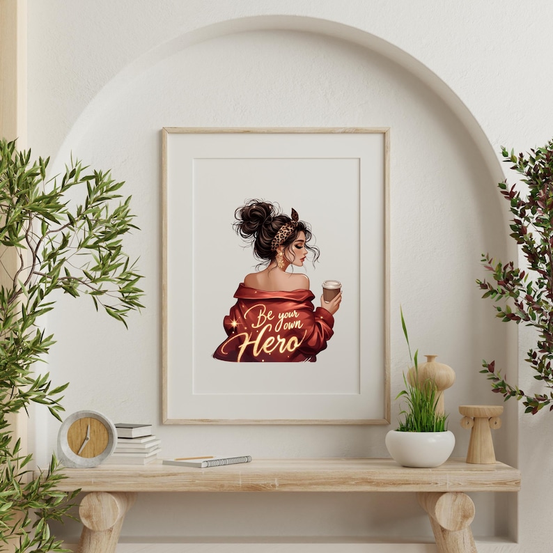 May include: Framed art print featuring a woman with dark hair in a bun, wearing a red jacket and holding a coffee cup. The print has the text "Be your own Hero" in gold script. The artwork is displayed on a wooden shelf.