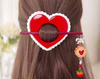 May include: A red heart-shaped hair accessory with a white scalloped border, secured by a dark red hair stick. A dangling charm hangs from the stick, featuring a small white bow, a round cookie with a red heart center, and a miniature strawberry.