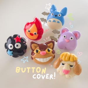 May include: A collection of colorful button covers featuring cartoon character designs. The covers include a blue creature, a red flame, a black cloud, a brown cat, a purple mouse, and a yellow dog. The text "BUTTON COVER!" is also visible.