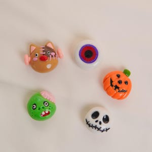 Puede incluir: Colección de cinco adornos decorativos con temática de Halloween. Incluye una criatura marrón con forma de gato, un globo ocular blanco con detalles rojos y morados, una calabaza naranja, un zombi verde y una calavera blanca.