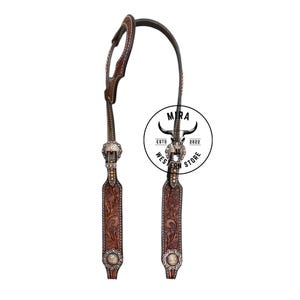 Può includere: Una testiera per cavallo in cuoio marrone scuro con dettagli floreali incisi, cuciture chiare e conchos in metallo argentato con incisioni floreali. Un logo circolare presenta un teschio di toro e il testo MIRA WESTERN STORE ESTD 2022.