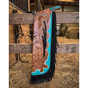 May include: Brown leather chaps with turquoise accents and black fringe. The chaps feature decorative tooling and three decorative squares with fringe. The chaps are hanging on a wooden fence.