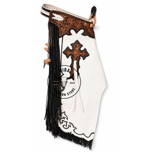 May include: White leather chaps featuring a brown tooled leather yoke with floral patterns, a large brown tooled cross appliqué, and long black fringe along the side. The waistband includes white lacing and a buckle. A logo with a skull is visible.