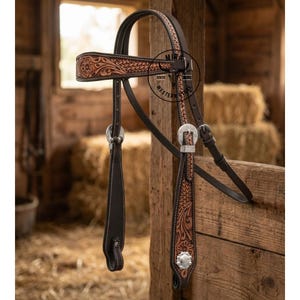 Può includere: Una briglia da cavallo in pelle marrone e beige con intricati disegni floreali e geometrici. La briglia presenta fibbie e dettagli argentati, con cuciture bianche. Lo sfondo mostra una struttura in legno rustica e balle di fieno.