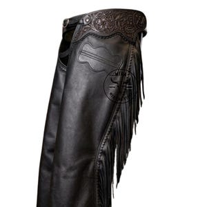 May include: Black leather chaps with decorative floral tooling on the waistband and a guitar-shaped design. The chaps feature long fringe along the outer leg seams, adding a classic Western flair. The leather appears to be smooth and supple.