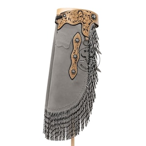 May include: Grey leather chaps featuring a tan leather yoke with intricate floral tooling and silver conchos. The side panel includes a vertical tan leather strip with silver studs and long, layered black and white fringe along the edge.