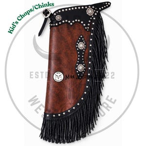 May include: A brown and black leather chaps piece for children featuring long black fringe, silver-toned metal studs along the edges, and decorative silver conchos. The item includes a black leather belt strap with a metal buckle and a logo.