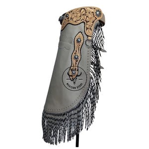 May include: A light gray leather chaps with intricate floral tooling on the upper portion. The chaps feature silver-toned studs and a long fringe along the sides. The image includes a logo that says "Western Store".