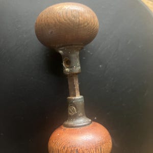 May include: A vintage wooden door knob with a metal spindle. The knob is brown and has a wood grain pattern. The spindle is silver and has a small metal plate with a design on it.