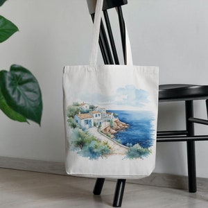 May include: White canvas tote bag with a watercolor illustration of a seaside village with blue buildings and a blue ocean. The bag has a black handle.