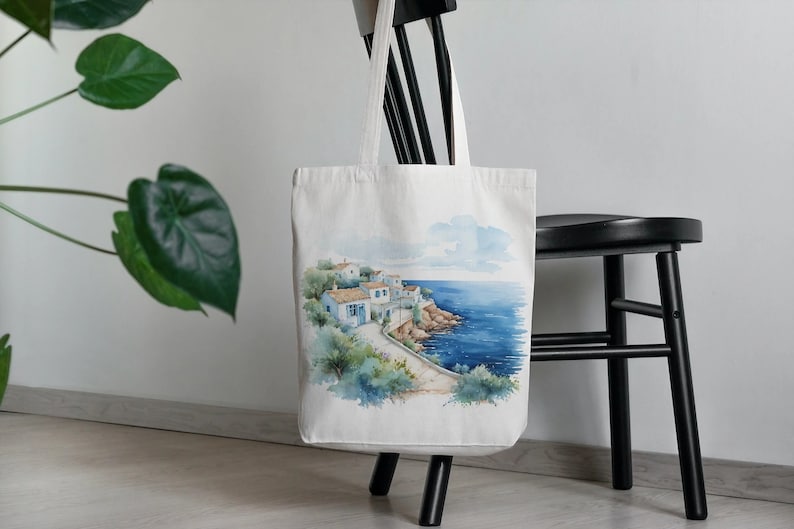 May include: White canvas tote bag with a watercolor illustration of a seaside village with blue buildings and a blue ocean. The bag has a black handle.