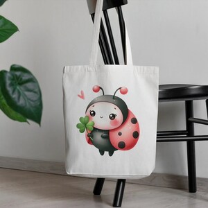 Puede incluir: Bolsa de tela blanca con una mariquita de dibujos animados sosteniendo un tr&eacute;bol de cuatro hojas. La mariquita tiene manchas negras y una concha roja.
