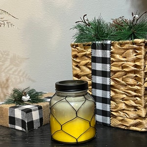 Pode incluir: Uma vela amarela num frasco de vidro com tampa preta e gaiola de arame. A vela está ao lado de uma cesta tecida com vegetação e uma caixa de presente embrulhada em serapilheira e tecido xadrez preto e branco. Os itens estão em uma superfície preta.
