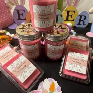 Pode incluir: Uma coleção de velas e ceras perfumadas cor-de-rosa com a etiqueta "Pink Sands" da L&C Homemade Candles. As velas estão em frascos de vidro com tampas de metal. Decorações de Páscoa, incluindo orelhas de coelho e recortes de ovos, no fundo.