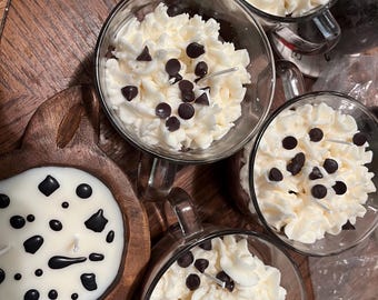 Könnte beinhalten: Vier gläserne Becher gefüllt mit weißer Zuckerglasur und Schokoladenstückchen, die an heiße Schokolade erinnern, und eine Holzschale mit einer weißen Kerze mit schwarzen Designs.