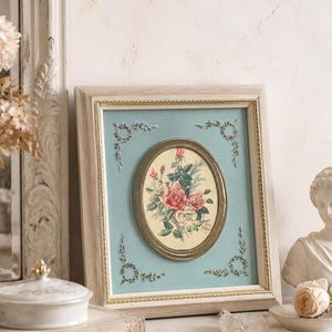 Puede incluir: Un estampado floral enmarcado con rosas rosas y hojas verdes. El estampado ovalado se encuentra sobre un fondo azul claro con detalles decorativos plateados. El marco es de madera clara con un borde interior dorado, creando una estética vintage.